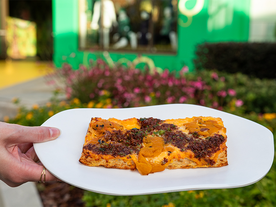 A hand holding a plate of the All Roads Lead to Oz Flatbread in front of Wicked: The Experience in Universal Studios Florida.
